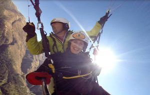 Dos personas con cascos en parapente se elevan sobre las majestuosas montañas de Granada. El cielo azul claro y la luz del sol crean una escena impresionante que captura a la perfección la esencia de "Vuelo en parapente en Granada". - ForTwo - Planes para parejas