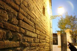 Al caer la noche, una pared de ladrillos bajo una farola brillante proyecta largas sombras sobre los árboles y las puertas, haciéndose eco del enigmático encanto del "Tour Conspiración en el Albaicín Granada", capturando el misterioso encanto de un paseo nocturno por el Albaicín. - ForTwo - Planes para parejas