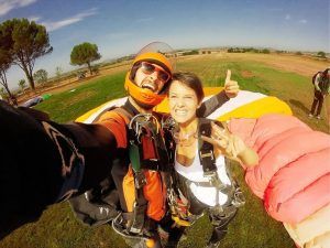 Después de un emocionante salto en paracaídas con BCN SkyDive cerca de Barcelona, dos personas con paracaídas posan con los pulgares hacia arriba y un símbolo de paz en un campo de hierba. - ForTwo - Planes para parejas