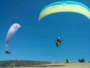 Dos personas experimentan un vuelo en parapente en Granada bajo un cielo azul claro, mientras otra captura la esencia impresionante de su vuelo desde tierra. - ForTwo - Planes para parejas