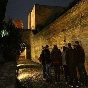 Un grupo de turistas se encuentra junto a un antiguo muro de piedra iluminado por la noche, inmersos en el misterio y el ambiente del "Tour Conspiración en el Albaicín Granada", con el encanto histórico del Albaicín como escenario de un edificio atemporal. - ForTwo - Planes para parejas