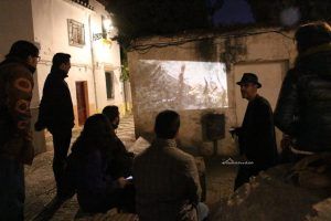 Los visitantes del Albaicín, Granada, quedan fascinados por una película proyectada sobre una antigua muralla, iluminada en la penumbra de la noche. La escena está llena de intriga, como si se tratara de una conspiración en desarrollo. Nombre del producto: Tour Conspiración en el Albaicín Granada. - ForTwo - Planes para parejas