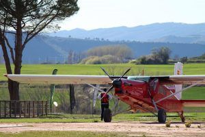 Junto a un avión de hélice rojo en un campo de hierba cerca de Barcelona, con montañas al fondo y una persona a su lado, esta escena es ideal para una aventura con "Paracaidismo en Barcelona desde 4000 metros - BCN SkyDive. - ForTwo - Planes para parejas