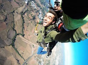 Dos personas saltan emocionantemente en paracaídas desde 4000 metros con BCN SkyDive, sobre campos y carreteras pintorescas, mientras el vibrante paisaje urbano de Barcelona se despliega maravillosamente en la distancia bajo un cielo azul claro. - ForTwo - Planes para parejas