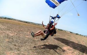 Dos personas se lanzan en paracaídas a un campo iluminado por el sol cerca de Barcelona, sonriendo y levantando los brazos después de un emocionante salto "Paracaidismo en Barcelona desde 4000 metros - BCN SkyDive". - ForTwo - Planes para parejas