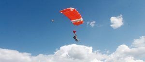 Dos personas saltan en paracaídas en tándem con un paracaídas rojo bajo un cielo azul brillante con nubes dispersas, encarnando la emoción del "Paracaidismo en Barcelona desde 4000 metros - BCN SkyDive. - ForTwo - Planes para parejas