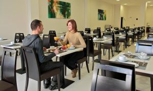 Dos personas disfrutan del desayuno y de una conversación en un moderno y sereno comedor, cuya interacción refleja la tranquilidad de la Habitación doble y circuito de aguas saludables en Balneario de Lanjarón (Granada). - ForTwo - Planes para parejas