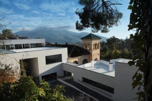 Este moderno edificio de fachada blanca y torre histórica se alza entre árboles y montañas bajo un cielo azul despejado. Cerca de allí, el famoso Balneario de Lanjarón ofrece la "Habitación doble y circuito de aguas saludables" para rejuvenecer y relajarse. - ForTwo - Planes para parejas