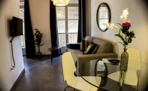 Apartamento para 2 en el centro de Granada cuenta con una moderna sala de estar con un elegante sofá gris, un televisor de pared y una mesa redonda de cristal con flores decorativas. Los ventanales iluminan el espacio con luz natural. - ForTwo - Planes para parejas