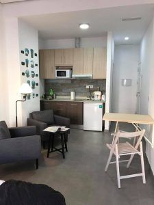 La encantadora cocina americana del Apartamento para 2 en el centro de Granada cuenta con armarios de madera, un cómodo sofá, una mesa pequeña, microondas, nevera y elegantes obras de arte. Ideal para una experiencia vibrante en el centro de Granada. - ForTwo - Planes para parejas