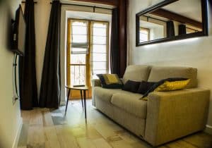 Apartamento para 2 en el centro de Granada cuenta con un sofá beige, una pequeña mesa redonda, una ventana grande y un televisor montado en la pared para una acogedora experiencia de sala de estar en el corazón de la ciudad. - ForTwo - Planes para parejas