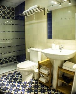 Un baño moderno en Apartamento para 2 en el centro de Granada cuenta con un suelo de baldosas geométricas, accesorios blancos, una ducha de cristal y estantes de madera llenos de toallas. - ForTwo - Planes para parejas