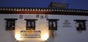 Un edificio blanco con retratos enmarcados y ventanas adornadas con flores al atardecer, con un cartel del Restaurante Estrellas de San Nicolás, ofrece la encantadora Cena para dos con vistas a la Alhambra. - ForTwo - Planes para parejas