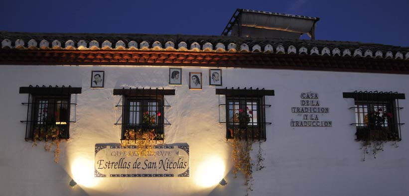 Un edificio blanco con retratos enmarcados y ventanas adornadas con flores al atardecer, con un cartel del Restaurante Estrellas de San Nicolás, ofrece la encantadora Cena para dos con vistas a la Alhambra. - ForTwo - Planes para parejas