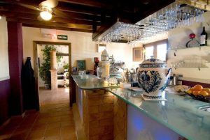 Un acogedor interior de cafetería con un mostrador de cristal, vasos colgantes y un jarrón de cerámica azul ofrece unas vistas encantadoras que recuerdan a la Cena para dos con vistas a la Alhambra en el restaurante Estrellas de San Nicolás a través de las ventanas y hacia otra habitación. - ForTwo - Planes para parejas