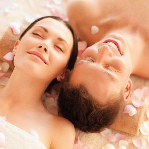 En Granada, una pareja disfruta del Masaje Mindfulness para 2 personas, relajándose sobre toallas rodeadas de pétalos de rosas rosas, con los ojos cerrados y sonriendo. - ForTwo - Planes para parejas
