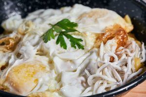 Primer plano de huevos fritos cubiertos con perejil, servidos sobre fideos blancos en una sartén moteada de negro, que recuerda al Menú Al Ándalus del Restaurante Carmen El Agua en Granada. - ForTwo - Planes para parejas