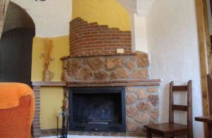 En una acogedora estancia al estilo de "Una noche en cueva romántica de turismo rural (Guadix)", una chimenea de piedra con chimenea amarilla y ladrillo realza la silla de madera y el sofá naranja, encarnando la encantadora esencia del turismo rural de Guadix. - ForTwo - Planes para parejas