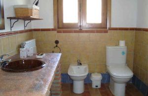 El encantador baño de la cueva romántica incluye lavabo, inodoro y bidé. Tiene paredes y suelos de baldosas, una ventana sobre el inodoro y una estantería con cesta, ideal para una auténtica experiencia rural en Guadix. - ForTwo - Planes para parejas