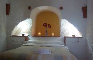 El acogedor dormitorio evoca el ambiente de una cueva romántica con una cama, tres velas encendidas en una estantería empotrada y una pared con acento amarillo. Captura a la perfección el encanto de Guadix para una experiencia de "Una noche en cueva romántica de turismo rural (Guadix)". - ForTwo - Planes para parejas