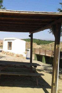 Una mesa de picnic a la sombra bajo un techo de madera rústica cerca de un edificio blanco y un cubo de basura añade encanto a esta zona rural, evocando la serenidad de "Dos noches en cueva romántica con hammam y masaje (Guadix). - ForTwo - Planes para parejas