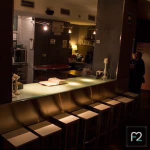 En el oscuro bar del Restaurante Ruta del Azafrán, hay taburetes alineados en el mostrador donde un hombre conversa en voz baja. La suave iluminación ambiental se suma a la atmósfera acogedora de su Degustación en Restaurante Ruta del Azafrán frente a la Alhambra. - ForTwo - Planes para parejas