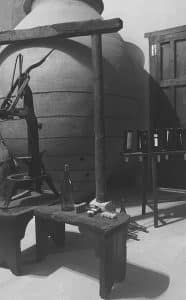 La fotografía en blanco y negro de una escena de bodega vintage en Bodega del Nero, que presenta una vasija de barro, una prensa de madera, botellas, corchos y un banco, evoca la tradición y el encanto de la elaboración del vino. Vívelo en vivo con Visita guiada a Bodega del Nero con tren turístico y degustación de 3 vinos. - ForTwo - Planes para parejas