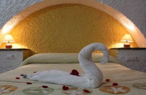 El encanto de una cueva romántica se captura con toallas en forma de cisne, pétalos de rosa sobre una cómoda cama y lámparas encendidas bajo un arco amarillo. Como nuestro "Dos noches en cueva romántica con hammam y masaje (Guadix). - ForTwo - Planes para parejas