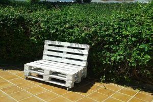 Un banco de paletas de madera blanca en un patio de baldosas, rodeado por un exuberante seto verde bajo un cielo soleado, crea un lugar tranquilo que recuerda a "Dos noches en cueva romántica con hammam y masaje (Guadix). - ForTwo - Planes para parejas