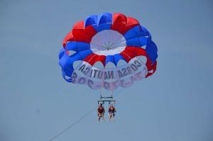 Dos personas experimentan "Hacer parasailing en Barcelona", su paracaídas rojo, blanco y azul crea un contraste impresionante contra el cielo despejado. - ForTwo - Planes para parejas
