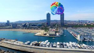 Experimente "Hacer parasailing en Barcelona" con impresionantes vistas de barcos amarrados, el vibrante paisaje urbano y un cielo azul claro. - ForTwo - Planes para parejas
