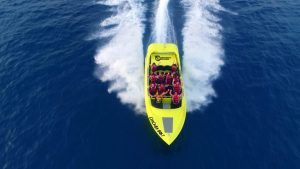 Vista aérea de un Chicken Boat amarillo brillante con pasajeros, "Surca las olas a toda velocidad en Chicken Boat en Barcelona", creando estelas de agua blanca en el océano azul profundo cerca de Barcelona. - ForTwo - Planes para parejas