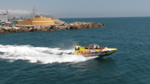 El Chicken Boat en Barcelona navega a toda velocidad cerca de un rompeolas en el vibrante océano azul, dejando un rastro de espumosas olas blancas a su paso. - ForTwo - Planes para parejas