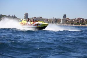 Una lancha rápida amarilla, similar a un Chicken Boat, avanza a toda velocidad por el agua frente al horizonte de Barcelona bajo un cielo azul brillante. - ForTwo - Planes para parejas