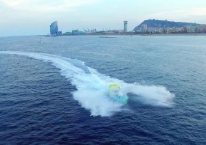 Un Chicken Boat, conocido como "Surca las olas a toda velocidad en Chicken Boat en Barcelona", corta el mar dejando una estela blanca cerca de la arquitectura moderna de Barcelona y las colinas distantes. - ForTwo - Planes para parejas