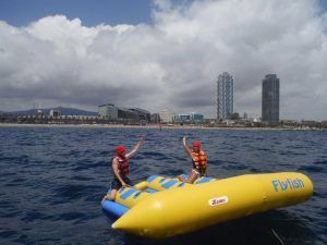 Dos amantes de las emociones fuertes disfrutan de la experiencia "Salta por los aires sobre el mar practicando el Flyfish en Barcelona", volando sobre las olas del océano con el horizonte de Barcelona y el cielo nublado como fondo pintoresco. - ForTwo - Planes para parejas
