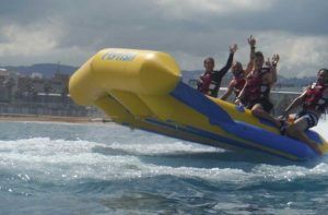 Un grupo de personas en una banana amarilla, sonriendo y levantando los brazos bajo un cielo nublado. Cerca de allí, "Salta por los aires sobre el mar practicando el Flyfish en Barcelona" crea una escena encantadora con el pintoresco mar de Barcelona como telón de fondo. - ForTwo - Planes para parejas