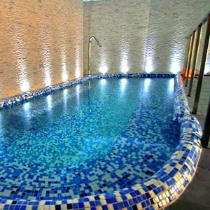 Experimente la tranquilidad en el Circuito en Spa + masaje en Granada Palace, donde una pequeña piscina cubierta con mosaicos azules está rodeada de paredes de piedra iluminadas, creando un ambiente sereno similar a un spa. - ForTwo - Planes para parejas