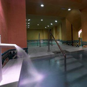 Un spa interior con paredes de azulejos, una fuente y escaleras que conducen a una piscina iluminada por luces cenitales. Mejore su experiencia con nuestra OFERTA: Circuito en Baños Árabes para 2 con cava, bombones y masaje opcional para una relajación total. - ForTwo - Planes para parejas