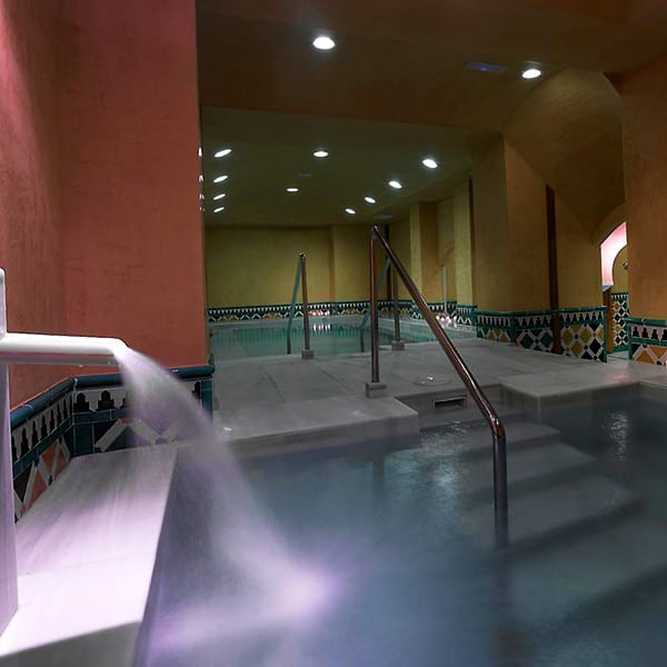 Un spa interior con paredes de azulejos, una fuente y escaleras que conducen a una piscina iluminada por luces cenitales. Mejore su experiencia con nuestra OFERTA: Circuito en Baños Árabes para 2 con cava, bombones y masaje opcional para una relajación total. - ForTwo - Planes para parejas