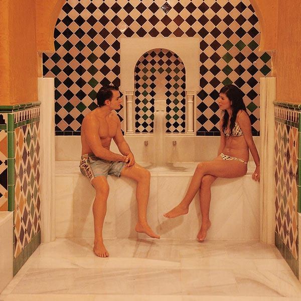 En traje de baño, dos personas se relajan en un banco de azulejos en una ornamentada sala con cava después de disfrutar de la OFERTA: Circuito en Baños Árabes para 2 con cava, bombones y masaje opcional, perfecta para relajarse. - ForTwo - Planes para parejas
