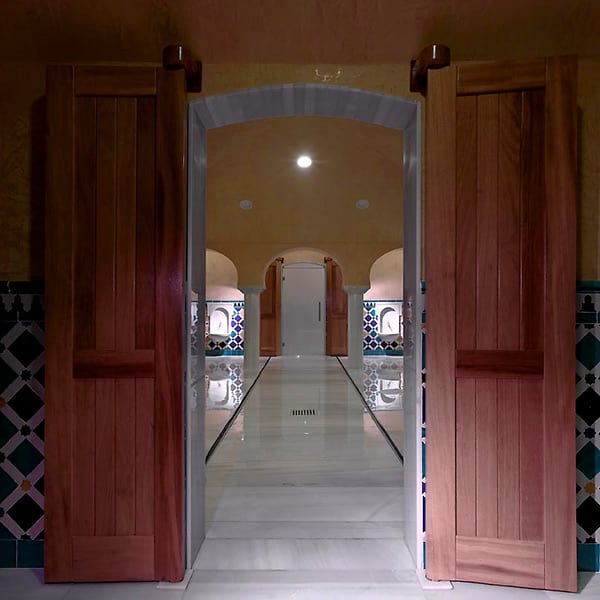 Puertas de madera conducen a un pasillo con arcos y paredes estampadas en una habitación con poca luz, que se asemeja a una OFERTA: Circuito en Baños Árabes para 2 con cava, bombones y masaje opcional. - ForTwo - Planes para parejas