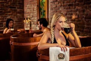 En Circuito Beer Spa de Granada, una mujer disfruta de una cerveza en una tina de madera junto a una pared de ladrillo, mientras otros se relajan cerca. Esta exclusiva experiencia Privado Premium está diseñada para la máxima relajación en pareja. - ForTwo - Planes para parejas