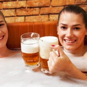 Dos personas sonríen y brindan con jarras de cerveza mientras disfrutan del Circuito Beer Spa Privado Premium para 2 personas en Granada, rodeado de una rústica pared de ladrillo. - ForTwo - Planes para parejas