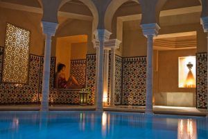 Una persona se relaja junto a una piscina en el Baño en Pareja Hammam Al Ándalus Málaga Baños Árabes de estilo marroquí, con azulejos y arcos ornamentados, suavemente iluminados por linternas, creando un escape tranquilo que hace eco del encanto del histórico Hammam Al Ándalus o de los tradicionales Baños Árabes. - ForTwo - Planes para parejas