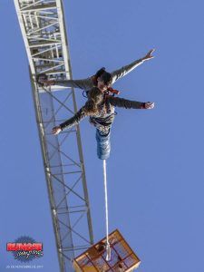 Una pareja atrevida abraza la emoción, con los brazos abiertos, mientras experimentan "Puenting en Barcelona en pareja", saltando en tándem desde una grúa contra el cielo azul claro de la ciudad. - ForTwo - Planes para parejas