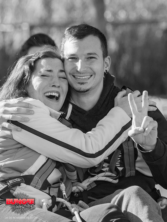 Una pareja feliz con arneses, sonriendo y disfrutando de su aventura de Puenting en Barcelona en pareja; una persona hace el signo de la paz. El logo de puenting en la esquina. - ForTwo - Planes para parejas
