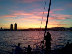 Figuras recortadas disfrutan de la puesta de sol a bordo de una "Puesta del sol en Barcelona desde Catamarán o velero", con el horizonte de Barcelona y el vibrante cielo nocturno como telón de fondo. - ForTwo - Planes para parejas