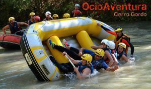 Los participantes con chalecos salvavidas y cascos se embarcan en un rafting grupal mientras el barco navega por las aguas bravas de Río Genil; Se ve el "Rafting en el Río Genil - Aguas Bravas" desde OcioAventura Cerro Gordo. - ForTwo - Planes para parejas