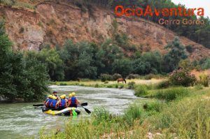 Personas haciendo rafting por el exuberante Río Genil mientras un caballo pasta cerca, con el texto "Rafting en el Río Genil - Aguas Bravas" arriba. - ForTwo - Planes para parejas